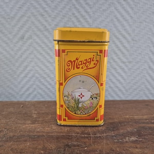 Op de afbeelding: Een rechthoekig geel metalen blik met het rode Maggi-logo aan de bovenzijde. Het midden toont een ronde illustratie van een witte soepterrine met een rood sterontwerp, omringd door een landschap met een windmolen, koeien en groen gebladerte.