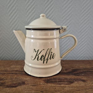 Op de afbeelding: Crème-kleurige emaille koffiepot met deksel, tuit en handvat. Het woord "Koffie" staat in een scriptlettertype op de voorkant. De pot heeft een zwarte rand en staat op een houten oppervlak.