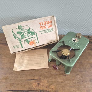 May include: A vintage green Turm 38 alcohol stove with a brass burner, a round dial, and a red adjustment knob. The stove is accompanied by its original cardboard box labeled Turm SK 38 and a printed instruction manual on a light tan paper sheet.