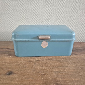 Op de afbeelding: Een vintage, rechthoekige, lichtblauwe metalen doos met een zilveren handgreep op het deksel. De doos heeft een licht versleten uiterlijk, met een kleine, ronde sticker op de voorkant. De doos staat op een houten oppervlak.