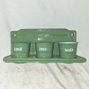 Op de afbeelding: Een groene metalen wandplank met drie geëmailleerde bakken. De bakken zijn gelabeld met "zand", "zeep" en "soda".