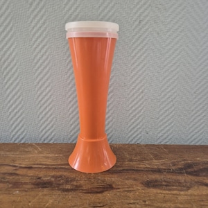 Può includere: Vaso di plastica arancione con bordo e coperchio bianchi. Il vaso ha una base svasata e un corpo conico e alto. Il vaso è appoggiato su una superficie di legno.