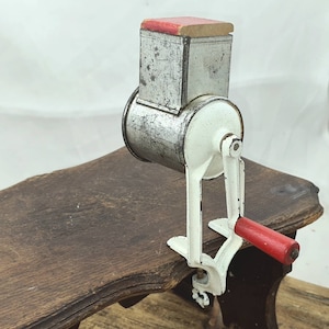 Puede incluir: Un molinillo de alimentos vintage blanco y plateado con una manivela roja. El molinillo está montado sobre una base de madera.