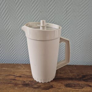 Può includere: Brocca in plastica beige chiaro con manico e coperchio. La brocca ha un beccuccio per versare e una superficie strutturata. Il manico è rettangolare e fissato al lato. Il coperchio ha un piccolo pezzo cilindrico al centro. Capacità di circa 2 litri.