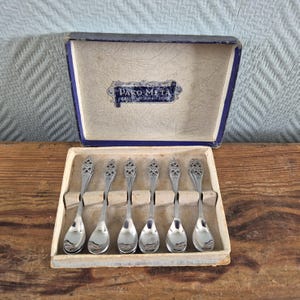 Puede incluir: Juego de seis cucharaditas de plata en una caja azul y blanca. La caja tiene el texto "Dako Meta" en ella. Las cucharas tienen un diseño adornado.