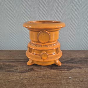 Op de afbeelding: Een oranje keramische vaas met een ronde boven- en onderkant, met drie kleine voetjes. De vaas heeft een cilindrisch lichaam met cirkelvormige en driehoekige decoratieve elementen. De kleur is een warm, aards oranje.