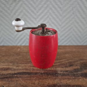 Op de afbeelding: Een vintage rode pepermolen. De houten behuizing is felrood en vertoont tekenen van slijtage. Hij heeft een metalen hendel met een witte keramische knop. Een klassiek keukengereedschap.