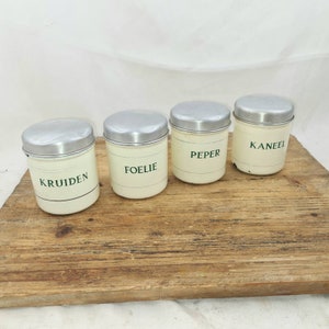 Op de afbeelding: Vier witte metalen kruidenpotten met zilveren deksels. De potten zijn gelabeld met "Kruiden", "Foelie", "Peper" en "Kaneel".