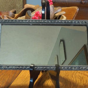 May include: A silver-toned vanity mirror with a decorative, ornate frame. The frame has a raised, rope-like design and two pearl-like accents on each side. The mirror is standing on a black stand.