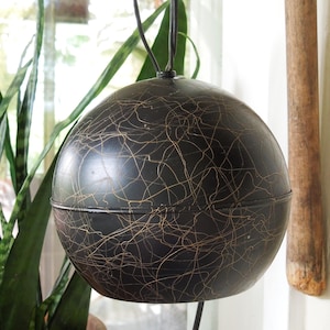May include: Black spherical pendant light with a gold, web-like pattern. The light hangs from a black cord, with a plant and wooden pole in the background. The light fixture is a decorative element.