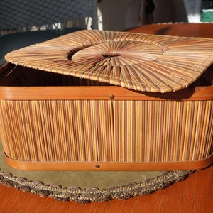 May include: Rectangular woven basket with a lid. The basket is made of light brown and tan reeds, with a darker brown wooden frame. The lid is partially open, revealing the interior. The basket sits on a green placemat with a decorative border.