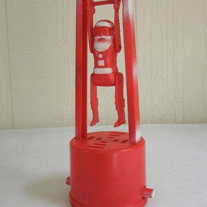 May include: A red plastic toy featuring a Santa Claus figure suspended from a horizontal bar between two vertical supports. The Santa figure has white painted details on the hat, beard, and belt. The entire structure is mounted on a round base.