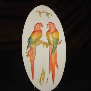 May include: A white oval ceramic wall plaque with a 3D design of two colorful parrots perched on a branch. The parrots have red, yellow, green, and blue feathers.