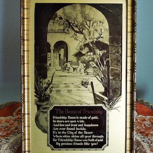 May include: A framed black and white print with a gold-colored frame. The print depicts an arched doorway leading to a courtyard with people. Text on the print reads "The House of Friendship" and a poem about friendship.