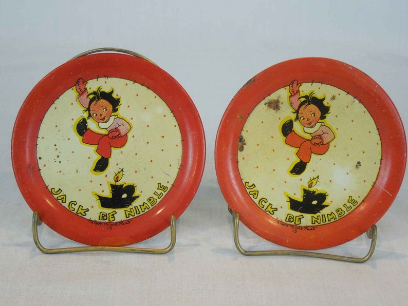 May include: Two red metal plates with a white center and a black and white illustration of a child jumping over a candle. The plates have the text "Jack Be Nimble" printed on them.