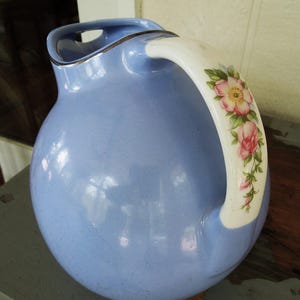 May include: A light blue ceramic pitcher with a round body and a curved handle. The handle is white with a floral design featuring pink roses and green leaves. The rim has a silver trim.