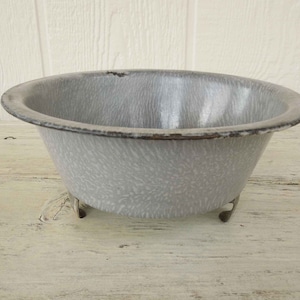 May include: A white enamelware bowl with a speckled pattern and a dark gray rim. The bowl has three small metal feet.