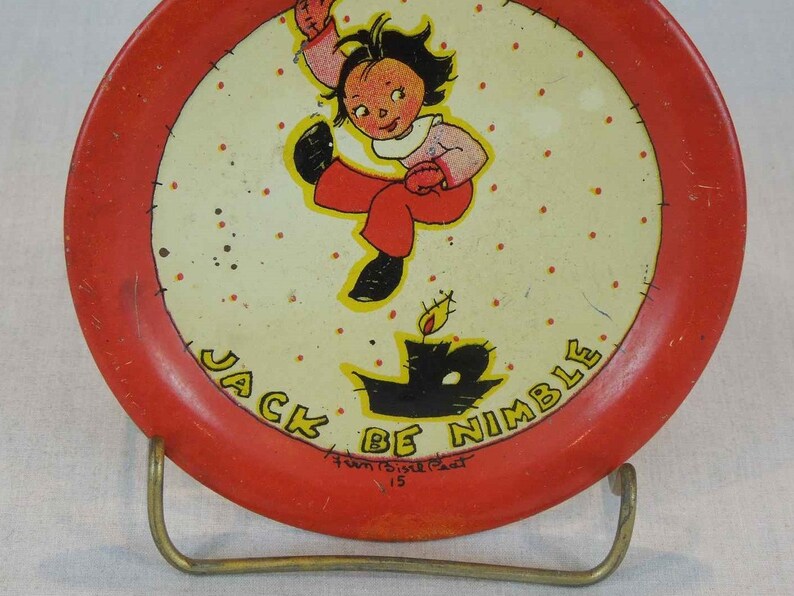 May include: A red and white metal plate with a cartoon image of a girl jumping over a candle. The plate has the text "Jack Be Nimble" around the edge and "Fun &  Fizzle Fact 15" at the bottom.
