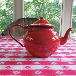 May include: A bright red enamel teapot with a rounded body and a curved spout. The teapot is sitting on a red and white checkered tablecloth.