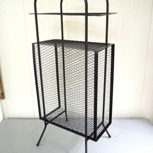 May include: A black metal storage rack featuring a rectangular frame with a wire mesh design. The unit includes two horizontal shelves and a tall, narrow vertical compartment with mesh sides, supported by four thin, angled metal legs.