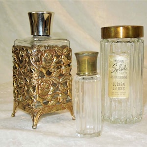 May include: Three vintage glass perfume bottles with gold and silver accents. The bottle on the left has a decorative gold rose design. The middle bottle has a gold cap. The bottle on the right has a gold cap and a label that reads "Tailspin Solid A Stick Cologne Lucien Lelong New York Chicago Net Wt 1.3 oz."