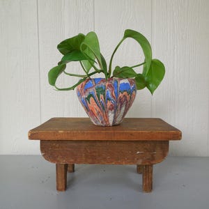 May include: A colorful, patterned planter with a green plant sits on a small, weathered wooden stool. The planter features a marbled design with shades of blue, orange, green, and white. The stool is a warm brown color and has four short legs.