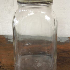 May include: A clear, rectangular glass jar with a metal lid. The jar has a wide mouth and a slightly curved body. The lid is a tarnished silver color. The jar is sitting on a wooden surface.