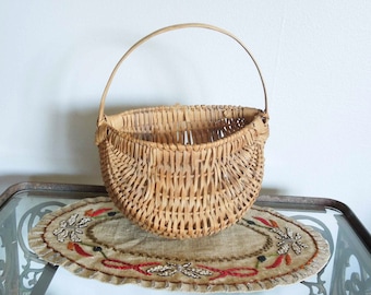 May include: A woven wicker basket with a single handle. The basket is oval shaped and has a natural brown color. The basket is sitting on a beige oval doily with red embroidery.