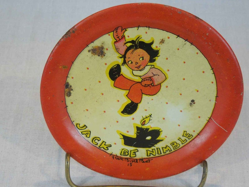 May include: A red and white metal plate with a cartoon illustration of a girl jumping over a black cat. The plate has a white background with red polka dots and the text "Jack Be Nimble" in black lettering. The text "Fun 15&cent;  Post 15" is also printed on the plate.