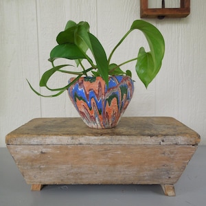 May include: A small, round planter featuring a marbled pattern of orange, blue, and green paint swirls. A vibrant green leafy plant grows from the top of the planter. The planter sits atop a rectangular, weathered wooden box with four small feet.