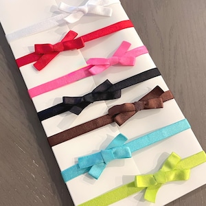 May include: A collection of colorful ribbon hair ties with small bow accents. The hair ties are in various colors, including white, red, pink, black, brown, teal, and lime green. The bows are attached to a white card.