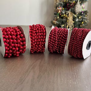 May include: Four spools of red beaded garland, perfect for holiday decorations. Each spool is tightly wound with small, round, shiny red beads. The spools are displayed on a wooden surface, with a decorated Christmas tree in the background.