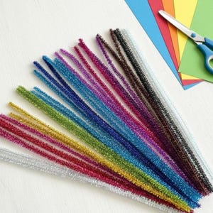 May include: A collection of multi-colored metallic pipe cleaners arranged in a fan shape, accompanied by a stack of rectangular construction paper in blue, red, orange, yellow, and green, and a pair of scissors with blue handles and silver blades.