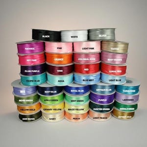 May include: A collection of spools of ribbon in various colors, including black, white, pink, and blue. Each spool is labeled with its color name, such as "Fuchsia," "Orange," and "Navy Blue." The ribbons are stacked neatly.