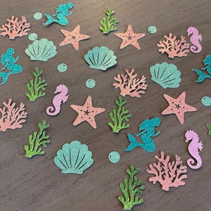 May include: Glittery mermaid-themed confetti in various shapes, including mermaids, seashells, starfish, seahorses, coral, and bubbles. The confetti features shades of teal, pink, and green, scattered on a wooden surface. Ideal for ocean-themed parties or crafts.