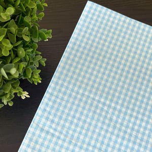 May include: A light blue and white gingham pattern fabric. The fabric is folded and placed on a dark brown surface. A green plant with small white flowers is in the upper left corner.