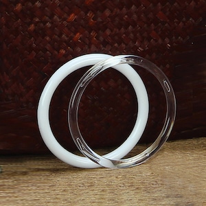 May include: Two bangle bracelets, one white and one clear, are stacked on top of each other. The bracelets are made of a smooth, hard material.