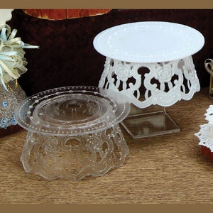 May include: Two clear plastic cake stands with a decorative lace design. The stand on the left is clear and the stand on the right is white.