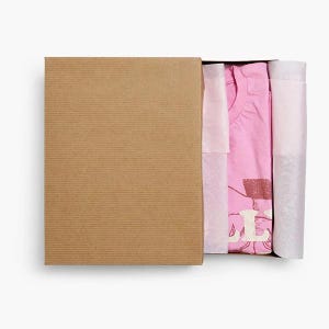 May include: A pink t-shirt folded inside a brown cardboard box, wrapped in white tissue paper. The shirt features a visible graphic print with the partial text "LL" in white lettering, partially obscured by the tissue paper packaging.