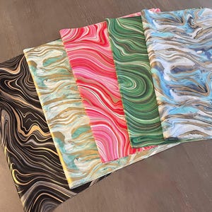 May include: A collection of colorful marbled paper sheets. The sheets display swirling patterns in black and gold, teal and gold, red and white, green and white, and blue and gold. The papers are arranged in a fan shape.