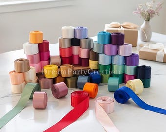 May include: A large collection of satin ribbon spools in a wide variety of colors including red, blue, pink, green, yellow, and purple. The spools are arranged in stacks and scattered across a white surface, with some ribbons unrolled.