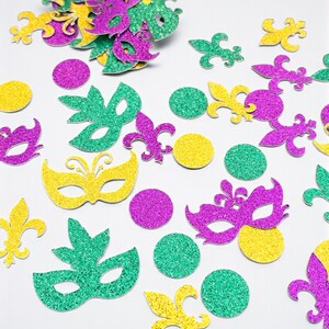 May include: Glittery Mardi Gras confetti in green, purple, and yellow. The confetti includes mask shapes, fleur-de-lis, and round circles. The festive decorations are scattered on a white surface.