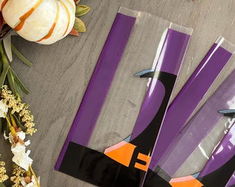 May include: Several cellophane bags with a purple border and a witch's leg design in black, orange, and blue. A small white and orange pumpkin and autumn foliage are in the background. The bags are on a grey surface.
