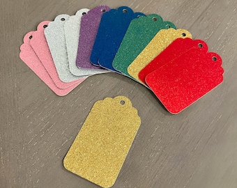 May include: A collection of glittery gift tags in various colors, including pink, silver, purple, blue, green, yellow, and red. Each tag has a scalloped edge and a hole for attaching a string or ribbon. One gold tag is separate.