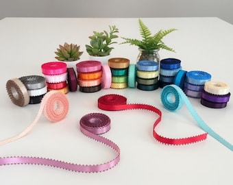 May include: Stacks of colorful satin ribbon spools in various shades, including pink, red, blue, and gold. Some spools are stacked, while others are unrolled, showcasing a decorative edge. Green plants in white pots and a glass vase are in the background.