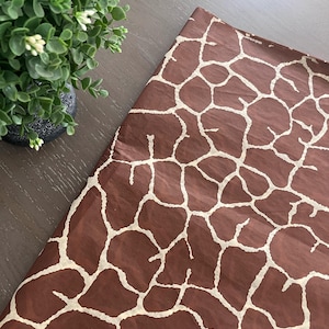 May include: Brown wrapping paper with a giraffe print pattern. The paper has a dark brown background with a network of irregular, off-white shapes resembling a giraffe's coat. A small green plant with white flowers is in the upper left corner.