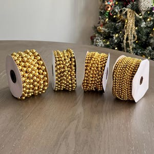 May include: Four spools of gold beaded garland sit on a wooden surface. The garland is made of small, round, shiny beads. A Christmas tree is visible in the background, suggesting holiday decor.