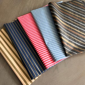 May include: Four folded sheets of patterned tissue paper arranged in a row. The patterns include tan with gold stripes, navy blue with gold stripes, bright red and white horizontal stripes, light blue and white stripes, and black with diagonal stripes.