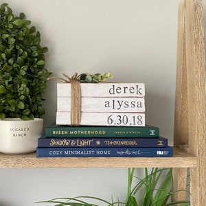 May include: A wooden shelf with a green wreath, a white candle, and a stack of books. The books are titled "Risen Motherhood", "Shadow & Light", and "Cozy Minimalist Home". The top book is a white stack of books with the names "derek", "alyssa", and "6.30.18" written on them.