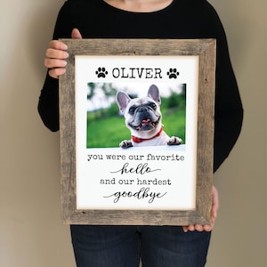 May include: A framed print with a photo of a dog and the text "Oliver" with paw prints on either side. The text below the photo reads "You were our favorite hello and our hardest goodbye".